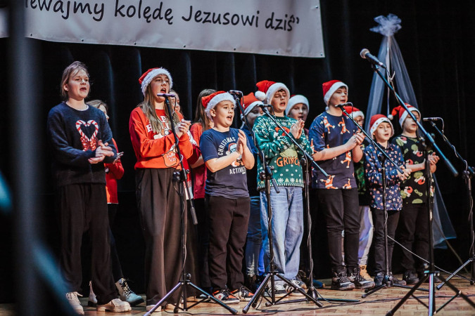 Uczniowie w mikołajkowych czapkach występują na scenie w trakcie Międzyszkolnego Konkursu Kolęd i Pastorałek, zdjęcie 2 z 5
